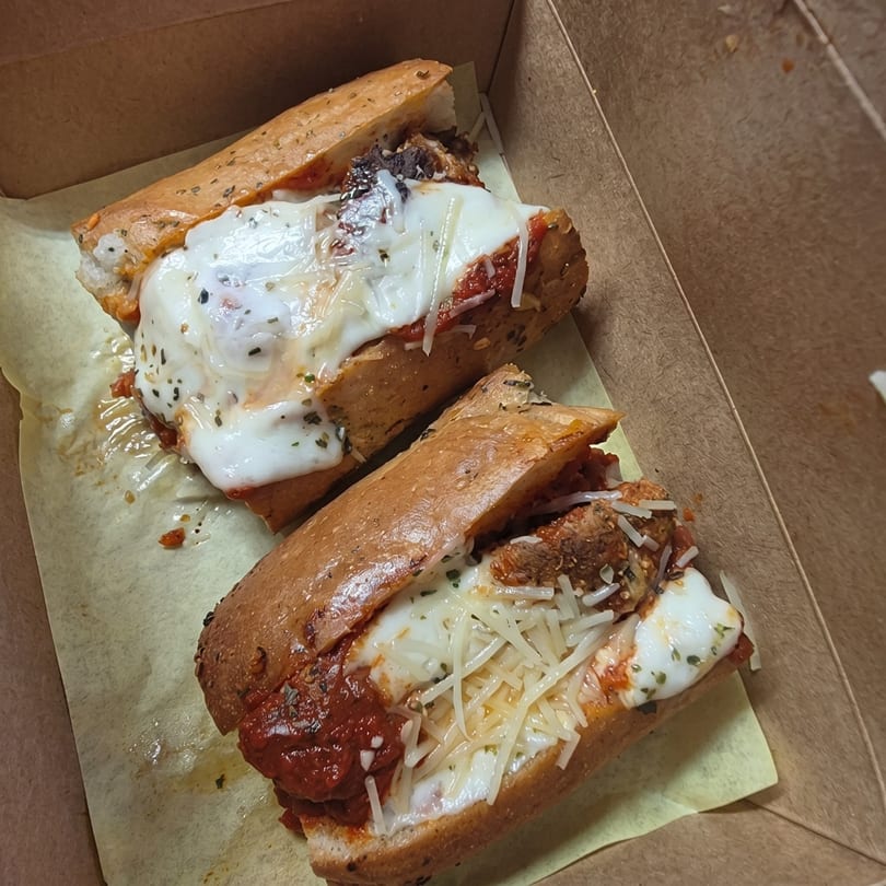 Two meatball subs with melted cheese and marinara sauce in a cardboard takeout box.