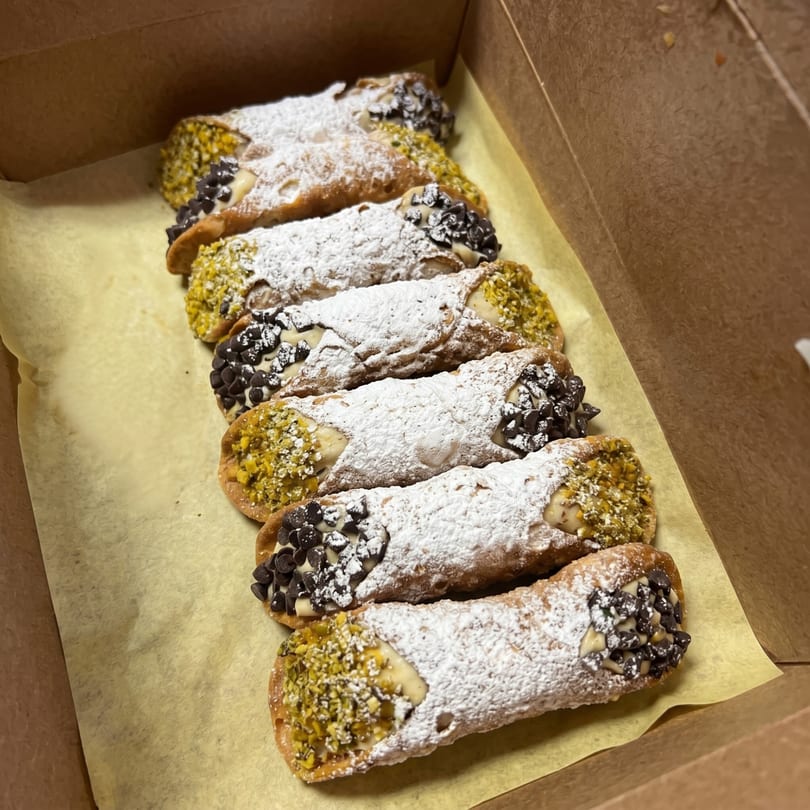 A row of cannolis, some with pistachios and some with chocolate chips, in a brown box.