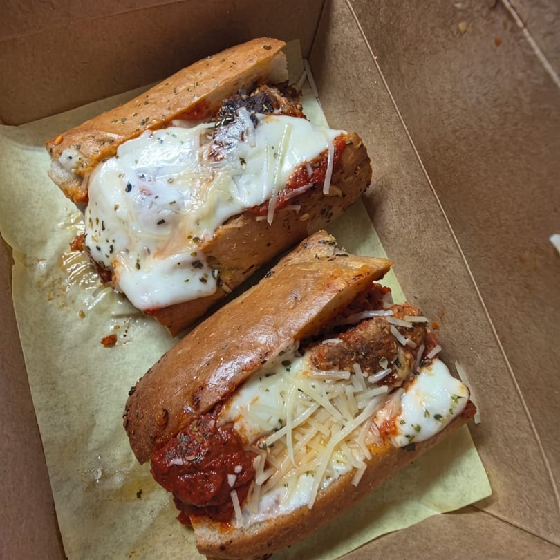 Two meatball subs with melted mozzarella and shredded Parmesan cheese in a cardboard takeout box.