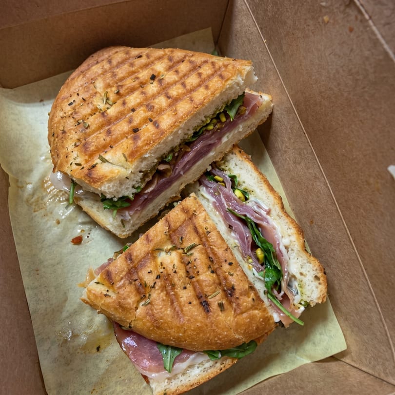 Two halves of a grilled panini sandwich with prosciutto, arugula, and cheese in a takeout box.