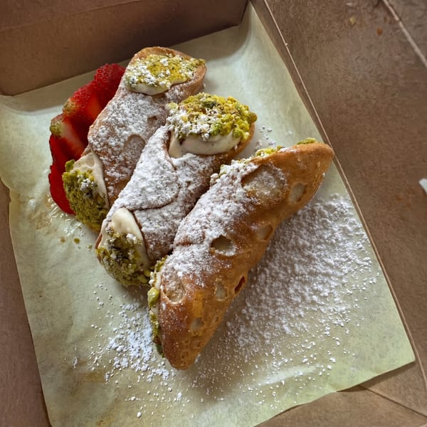 Three cannoli with powdered sugar and pistachio, with a sliced strawberry, in a takeout box.