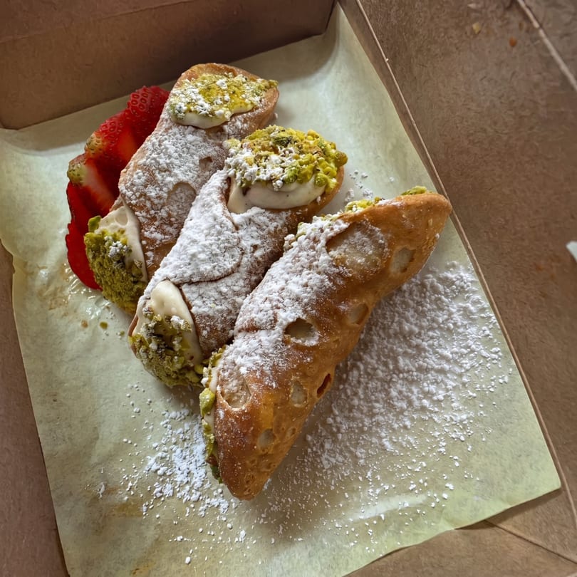 Three cannoli with powdered sugar and pistachio, with a sliced strawberry, in a takeout box.