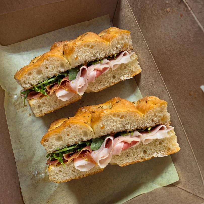 Two focaccia sandwiches with prosciutto, ham, and arugula, in a brown takeout box.